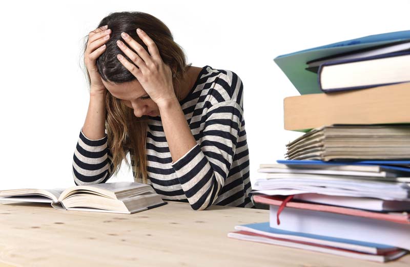 Young stressed student girl studying pile of books on library desk preparing MBA test or exam in stress feeling tired and overwhelmed in youth education concept